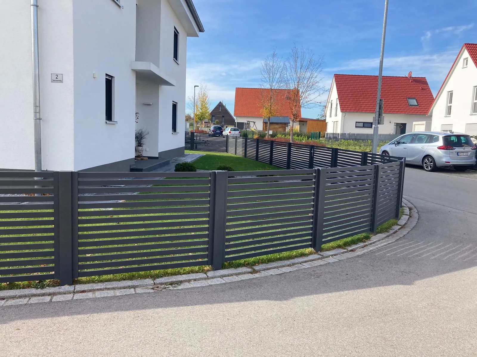 Dunkler Aluminiumzaun mit waagerechten Lamellen entlang eines hellen Hauses und einer Straße.